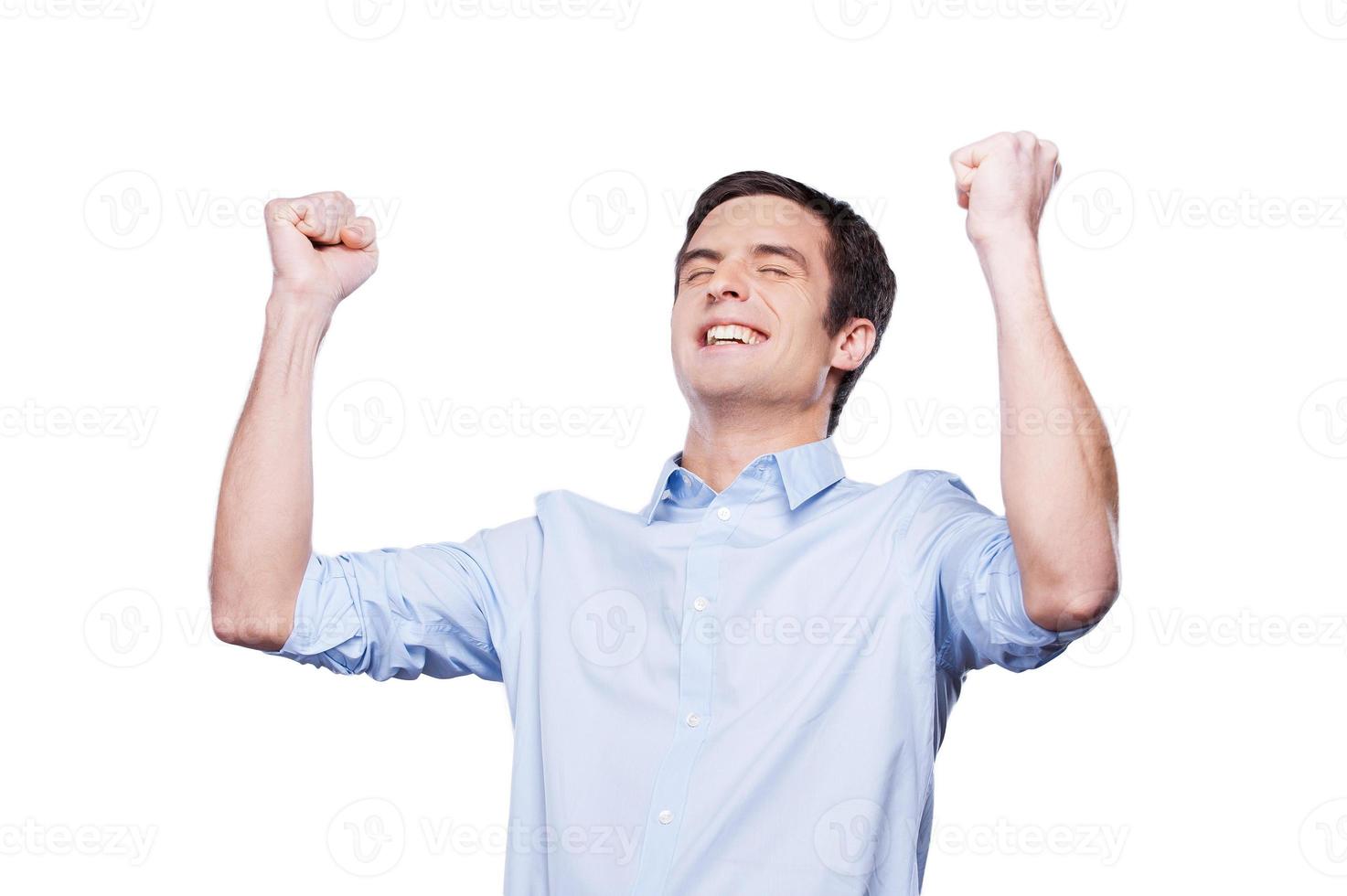 qué éxito feliz joven con camisa azul gesticulando y manteniendo los ojos cerrados mientras está de pie aislado en blanco foto