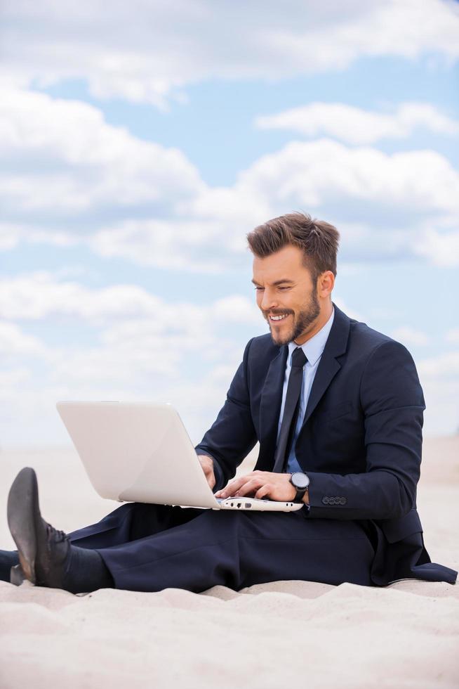 He found a peaceful place to work. Handsome young man in formalwear working on laptop and smiling while sitting on sand in desert photo