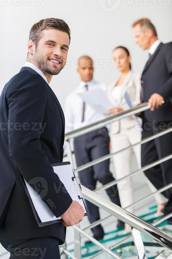 Young and successful. Low angle view of confident young man in formalwear looking over shoulder and smiling while moving up by staircase with people in the background photo