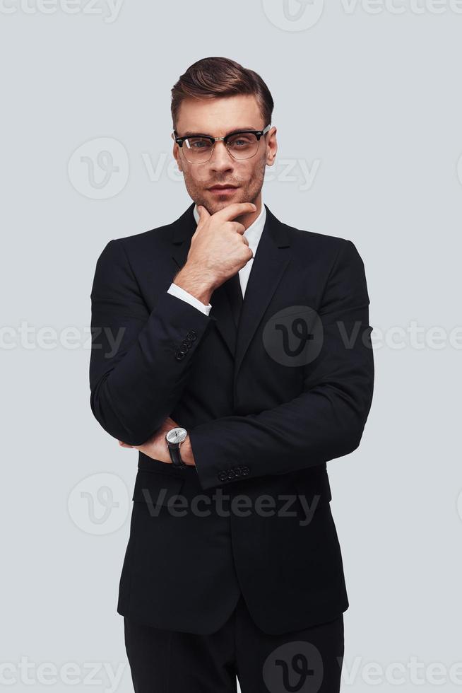 Lost in thoughts. Serious young man in full suit keeping hand on chin and looking at camera while standing against grey background photo