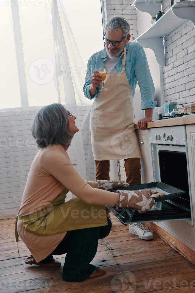 alegre pareja mayor en delantales preparando una cena saludable en el horno y sonriendo mientras pasa tiempo en casa foto