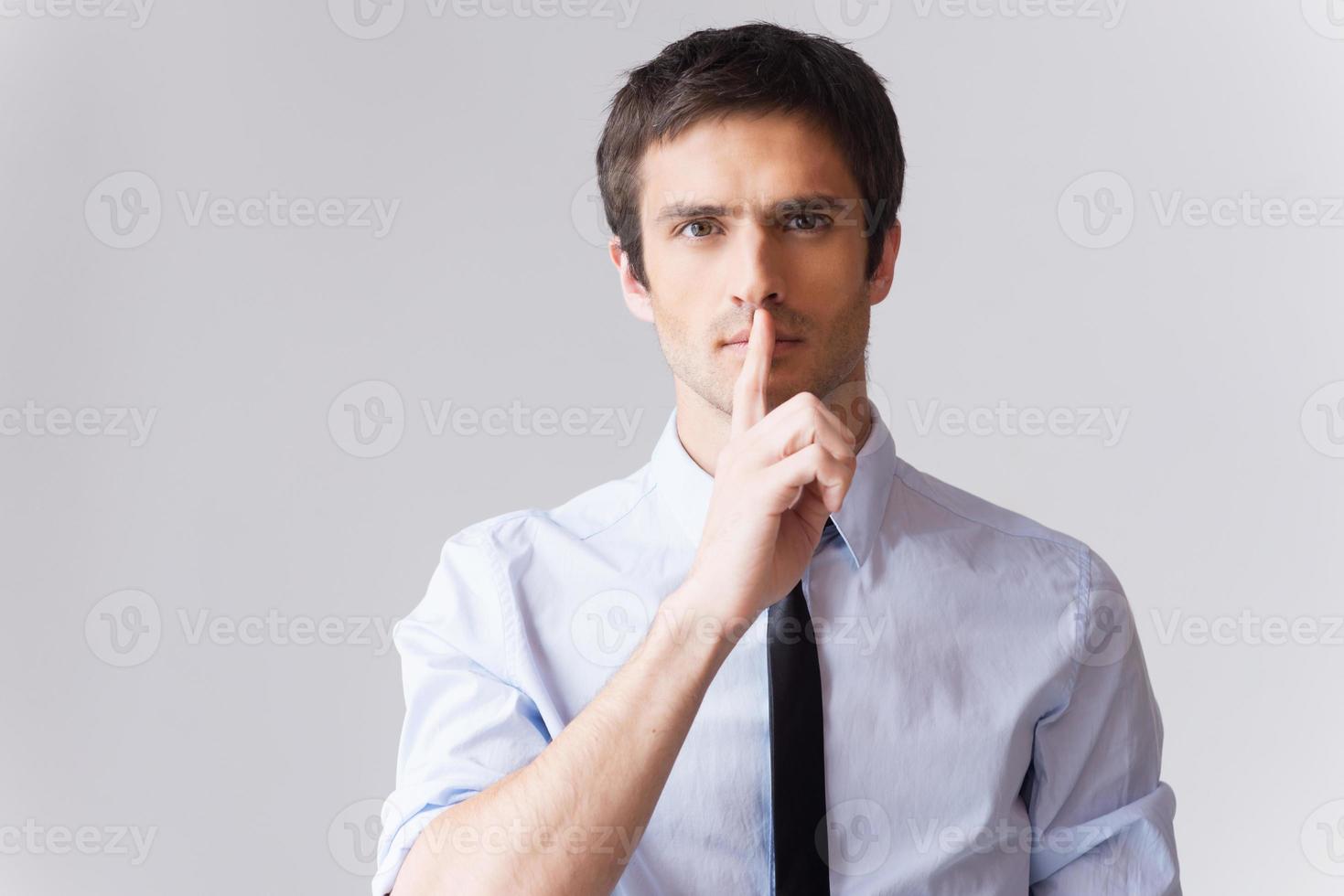 Keep a secret Confident young man in white shirt looking at camera and holding finger on lips while standing against grey background photo