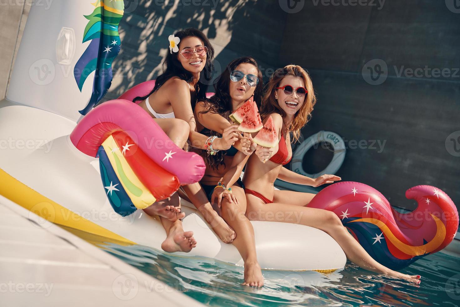 Attractive young women in swimwear smiling and eating watermelon while floating on inflatable unicorn in swimming pool outdoors photo