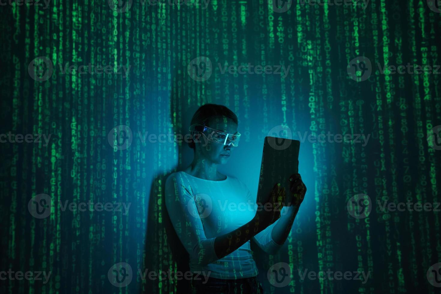 Confident woman in futuristic glasses using digital tablet against dark background with green symbols photo