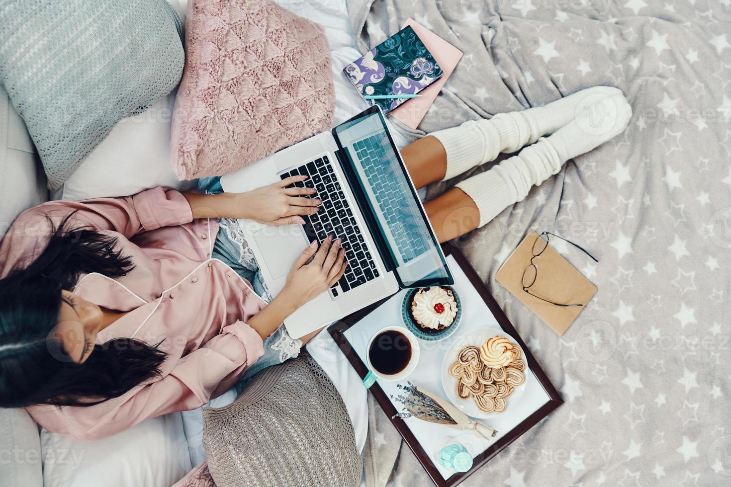 Top view of beautiful young woman in pajamas using laptop while resting in bed at home photo