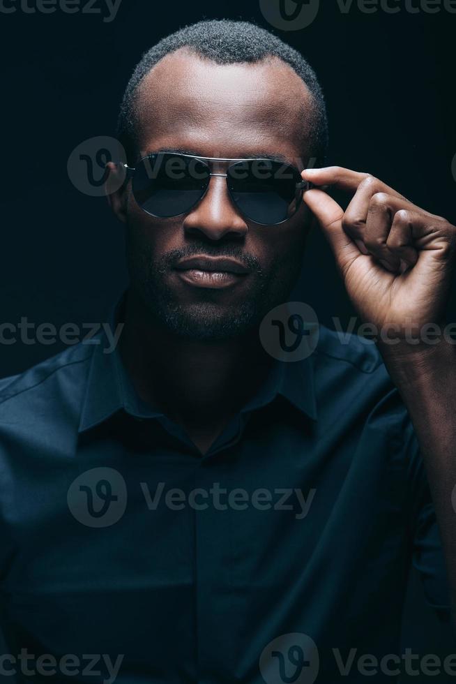 fresco y de moda. retrato de un apuesto joven africano ajustando sus gafas de sol mientras está frente a un fondo negro foto