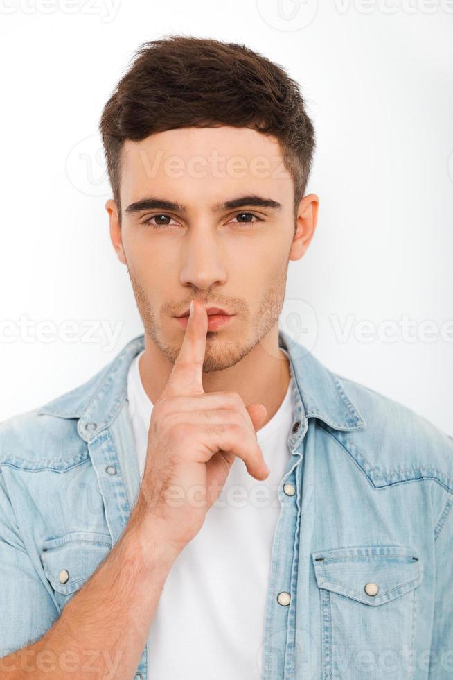 Keep my secret Serious young man looking at camera and holding finger on lips while standing against white background photo