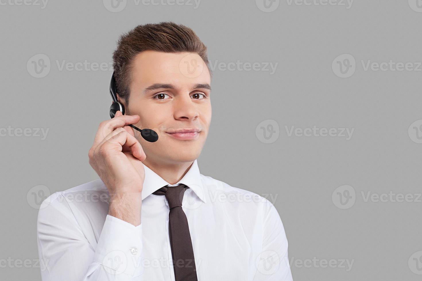 Man in headset. Cheerful young man in formalwear adjusting his headset and smiling at camera while standing against grey background photo