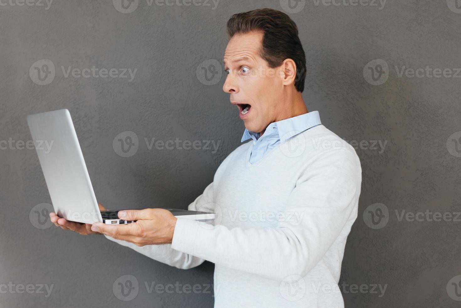 What is that Surprised mature man looking at laptop and keeping mouth open while standing against grey background photo