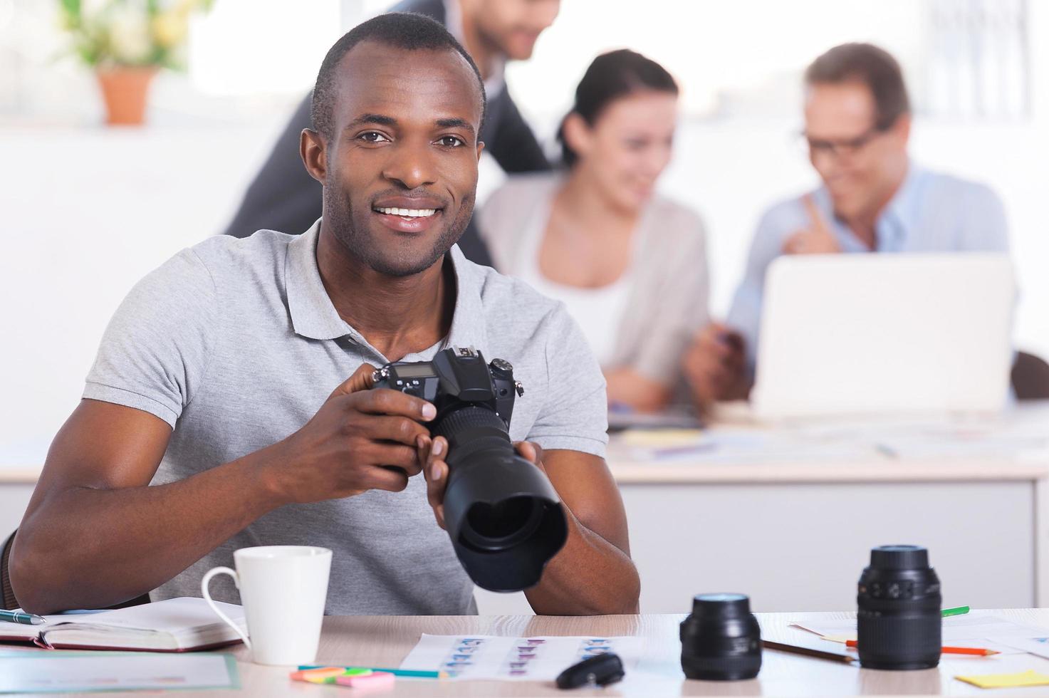 Creative people at work. Handsome young African man holding camera and smiling while three people working on background photo