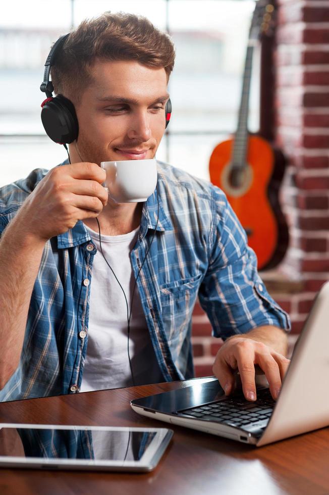 Relaxing with his favorite music. Handsome young man in headphones working on laptop and drinking coffee while acoustic guitar laying in the background photo