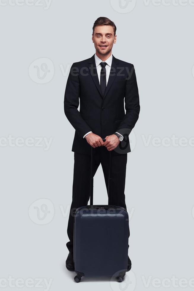 Business travel. Full length of good looking young man with suitcase smiling and looking at camera while standing against grey background photo