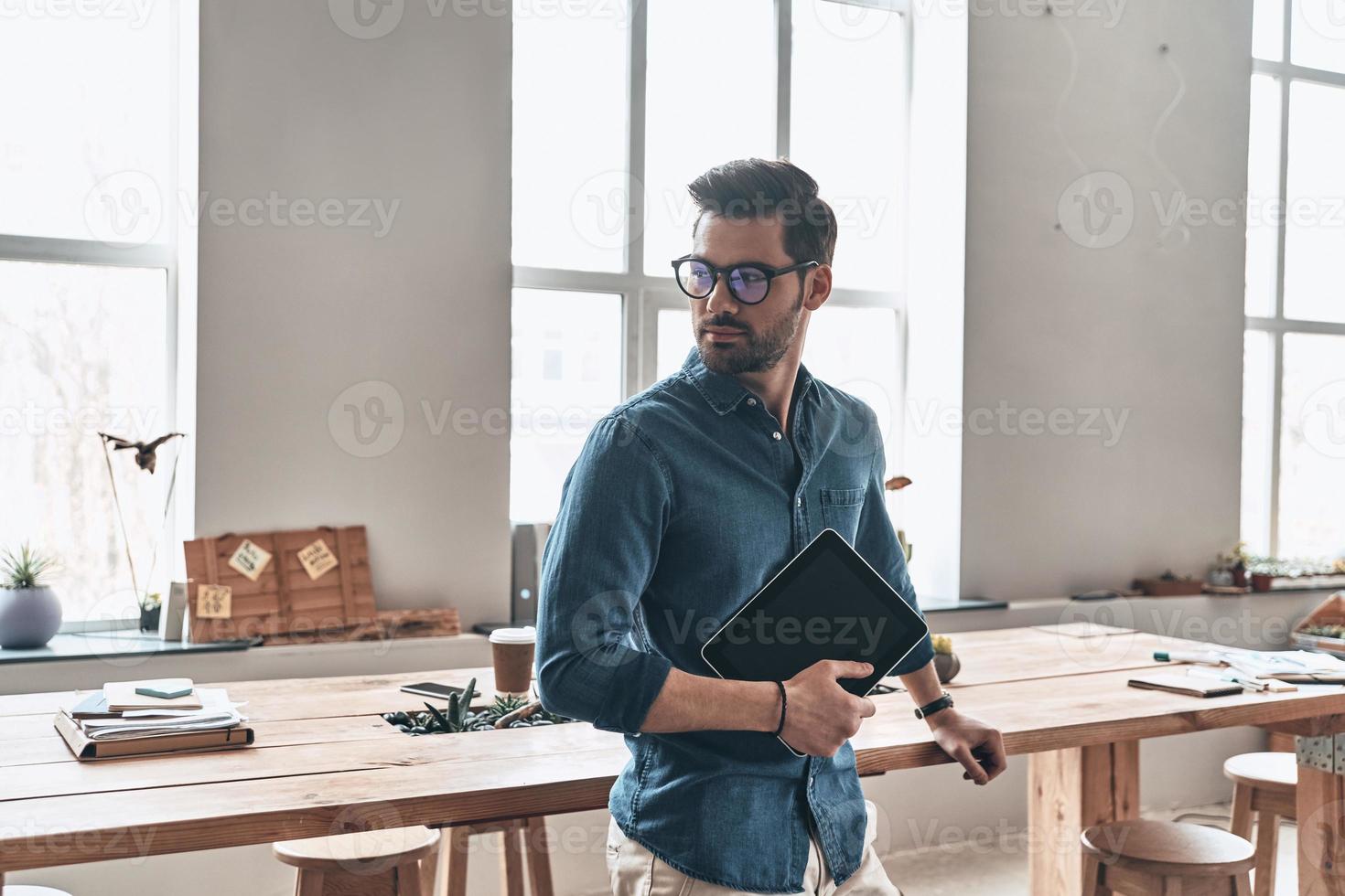 Confident and handsome. Handsome young man holding digital tablet and looking away while standing in the office photo