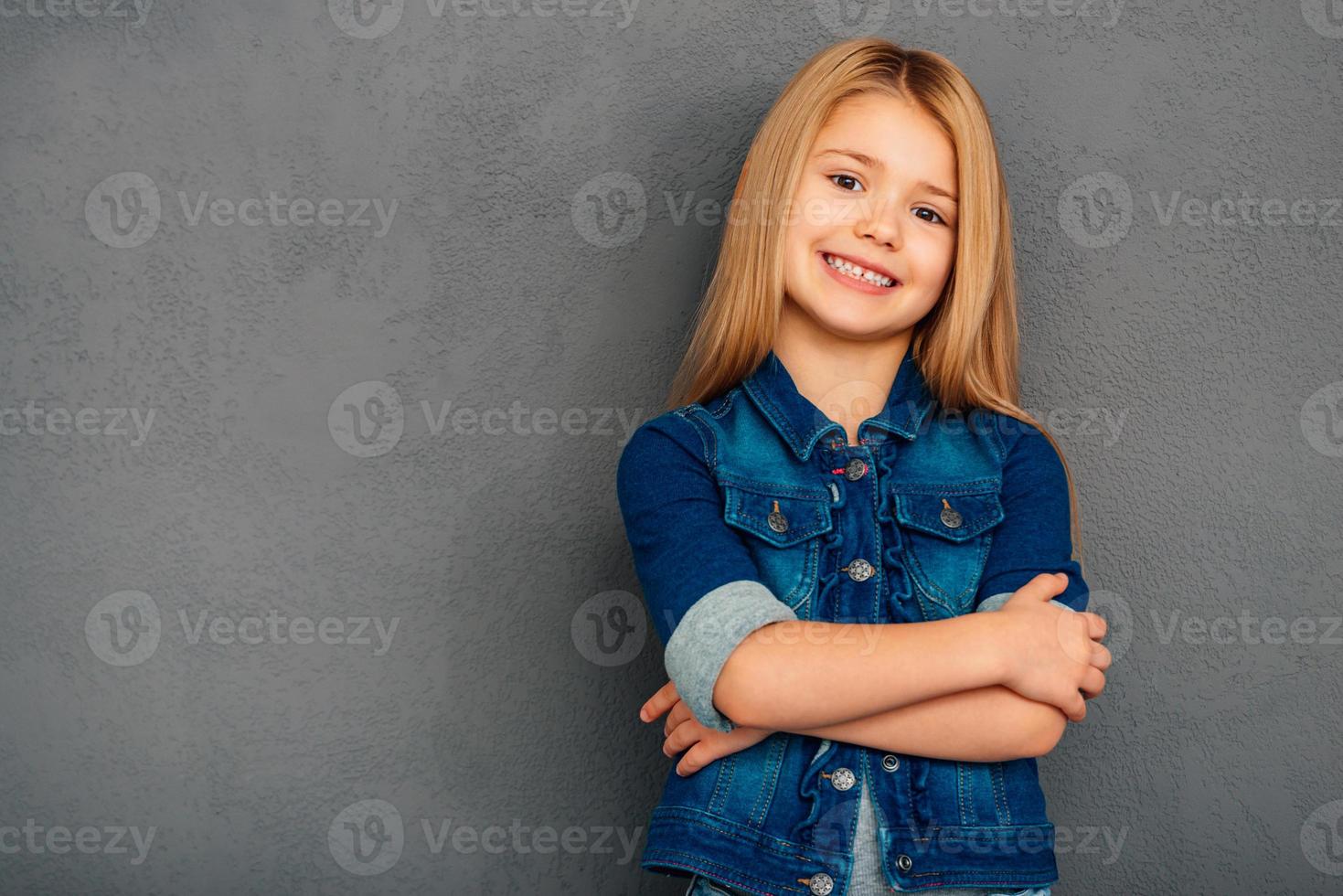 Little cutie. Cheerful little girl holding arms crossed and looking at camera with smile while standing against grey background photo