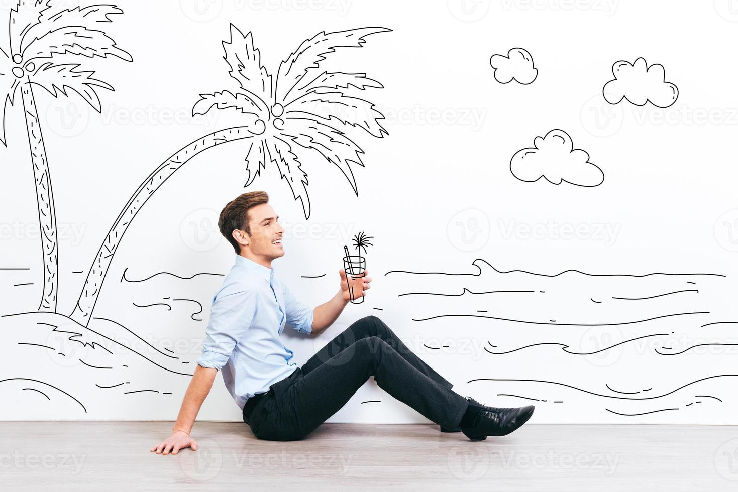 Dreaming about vacation. Young handsome man holding drawn cocktail and looking away with smile while sitting on the floor with illustration of resort in the background photo