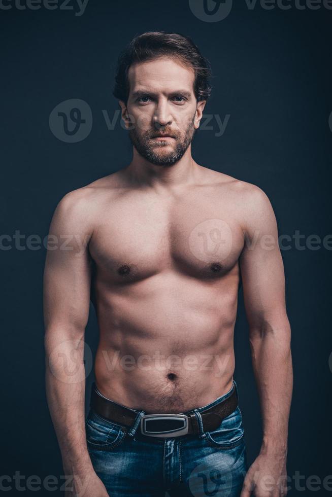 Strong and confident. Young shirtless man with serious face looking at camera while standing against black background photo
