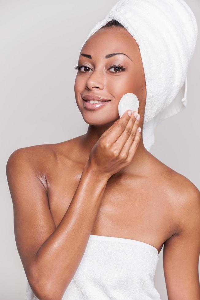 Tratamiento facial. retrato de una alegre joven afroamericana sin camisa sonriendo a la cámara y tocando la cara mientras se enfrenta a un fondo gris foto