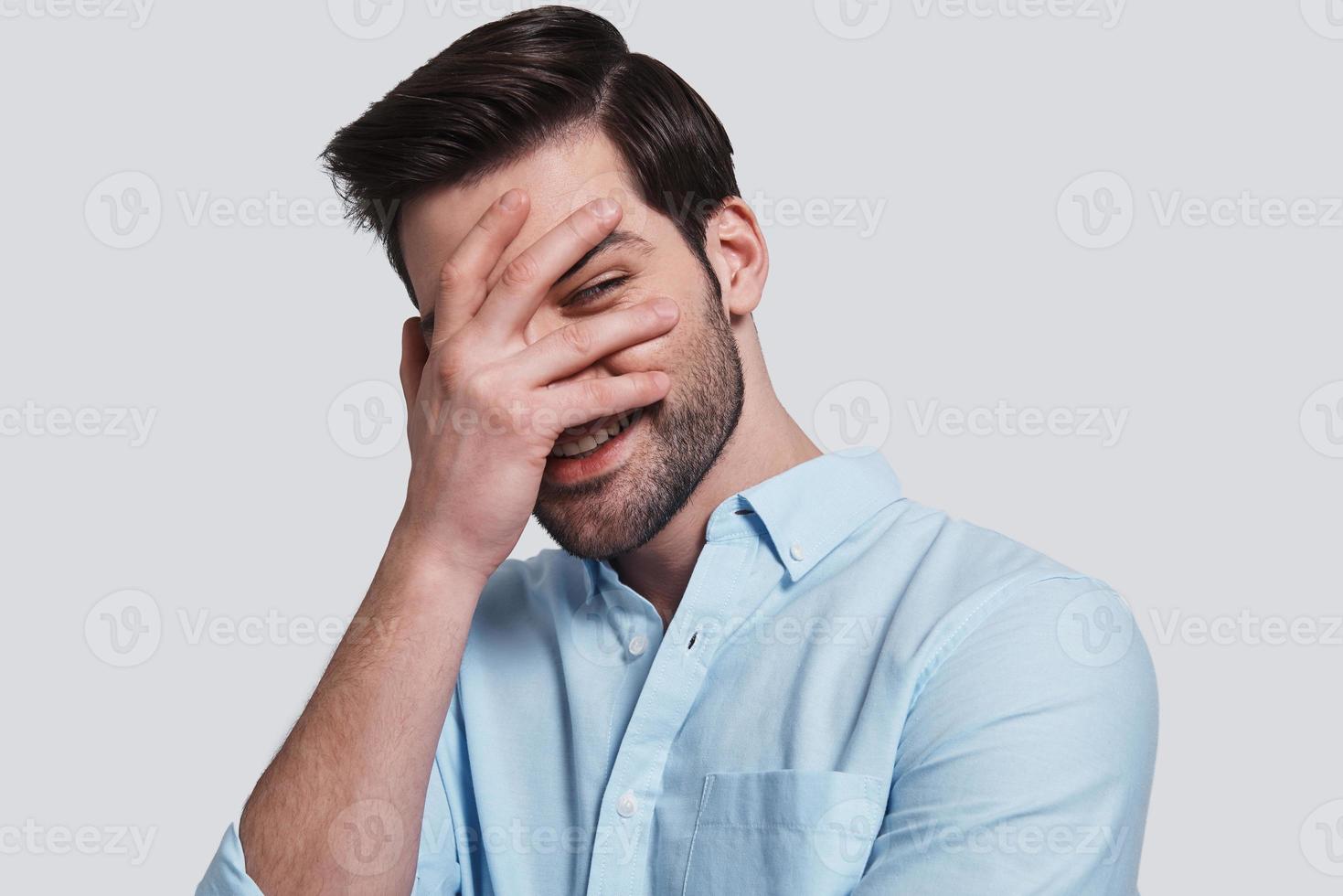 Feeling curious.  Handsome young man covering eyes with hand and peeking while standing grey background photo