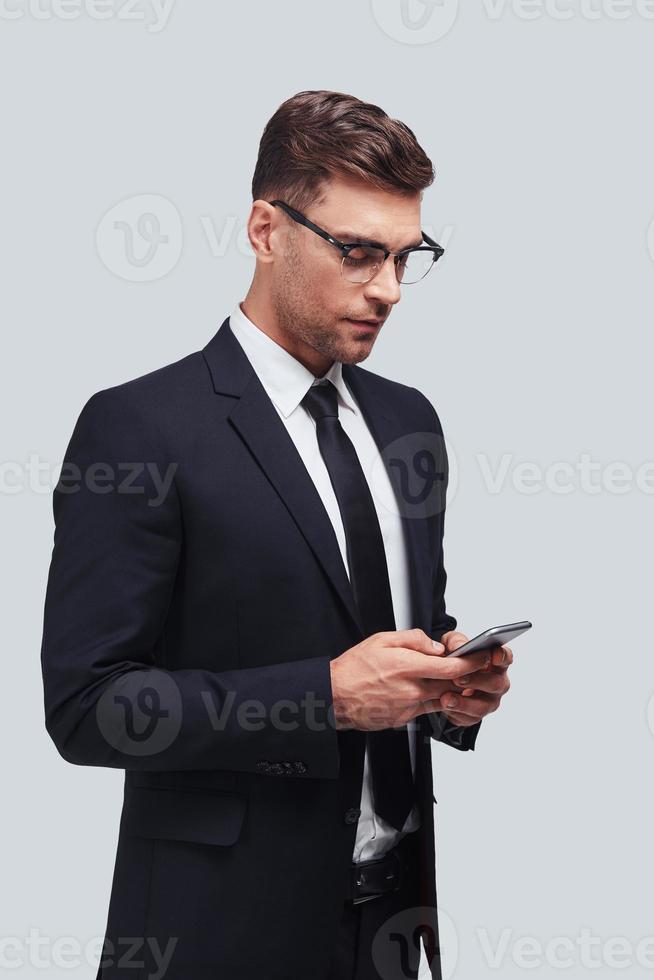 Another business message. Handsome young man using his smart phone while standing against grey background photo