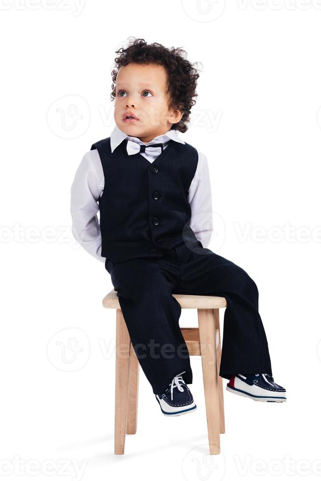 Me sentaré aquí y no haré nada, un niñito africano mirando hacia otro lado mientras estoy sentado en un taburete con fondo blanco. foto