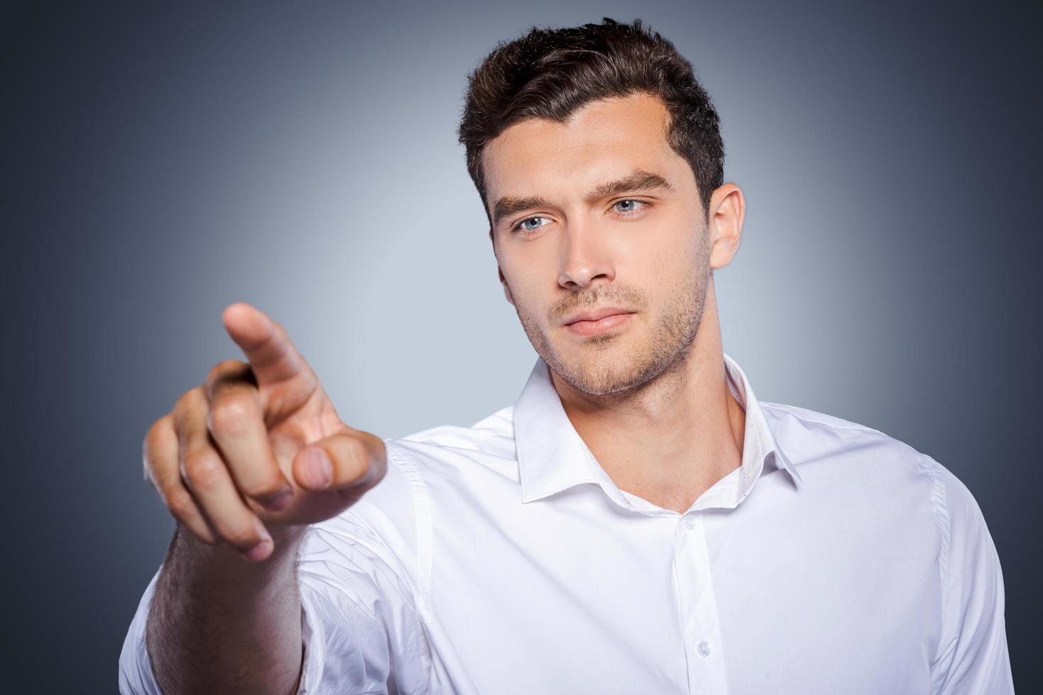 Working on transparent wipe board. Handsome young man in white shirt touching transparent wipe board while standing against grey background photo