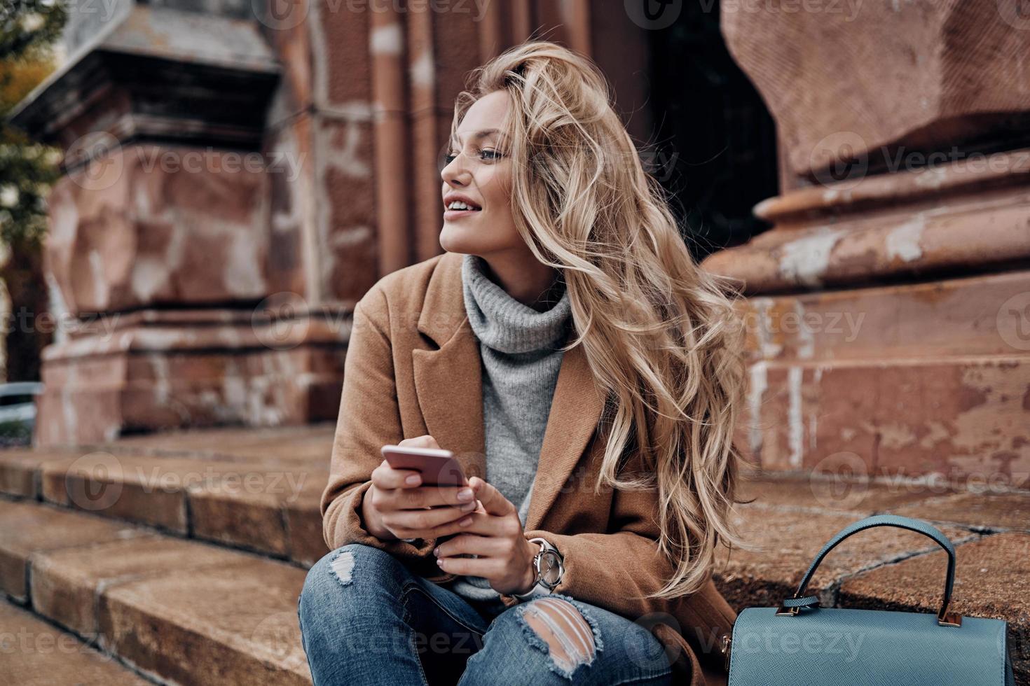 Thoughtful beauty. Beautiful young woman holding her smart phone and looking away with smile while sitting on stairs outdoors photo