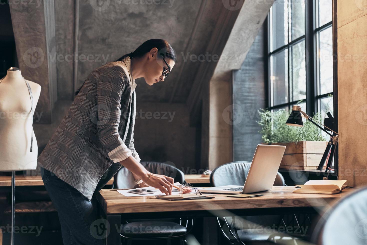 Achieving success as a designer. Concentrated young woman in eyewear looking at sketches while standing in her workshop photo