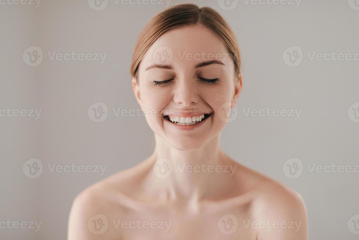 Sincere smile. Portrait of gorgeous young woman with freckles on face smiling and keeping eyes closed while standing against grey background photo