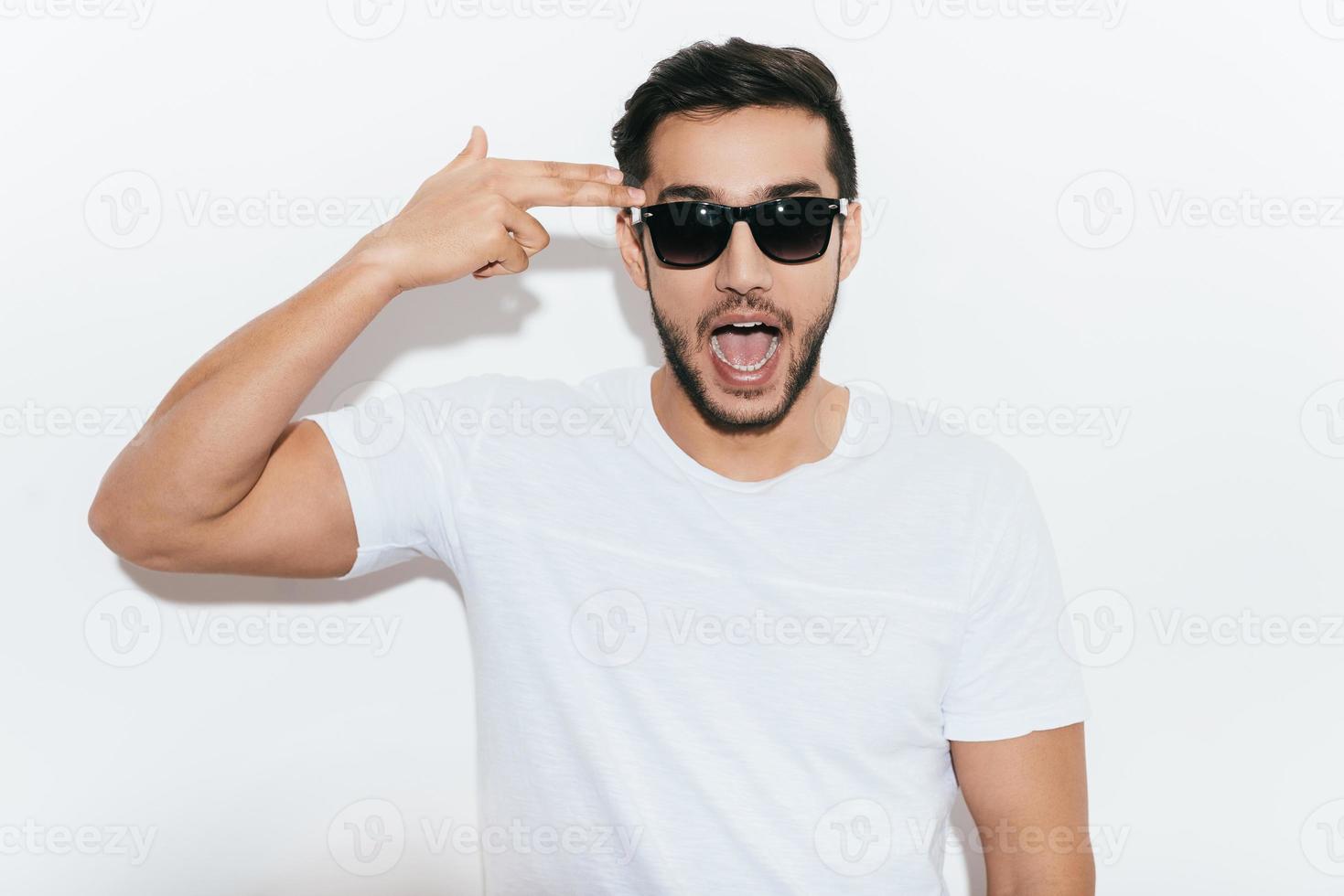 Bang Playful young Indian man in sunglasses gesturing handgun near head and making a face while standing against white background photo