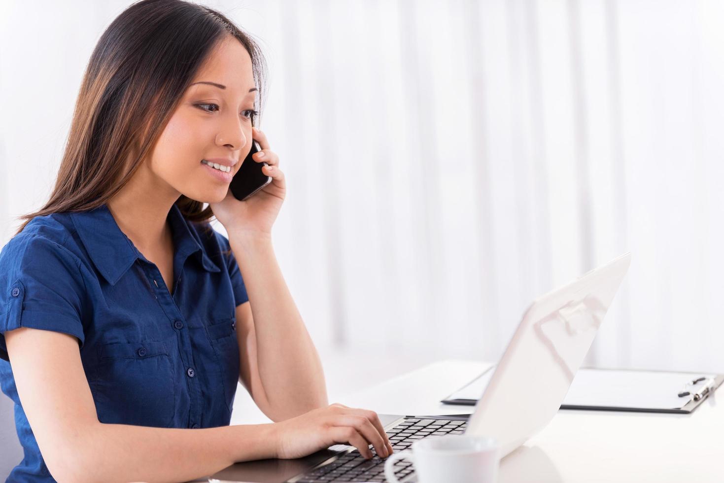 Discussing new project. Side view of beautiful young Asian woman talking on the mobile phone and looking at laptop while sitting at her working place photo