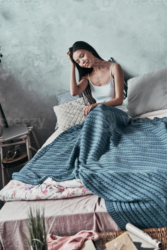 Just one more minute in bed. Attractive young woman keeping hand in hair and smiling while sitting on the bed at home photo