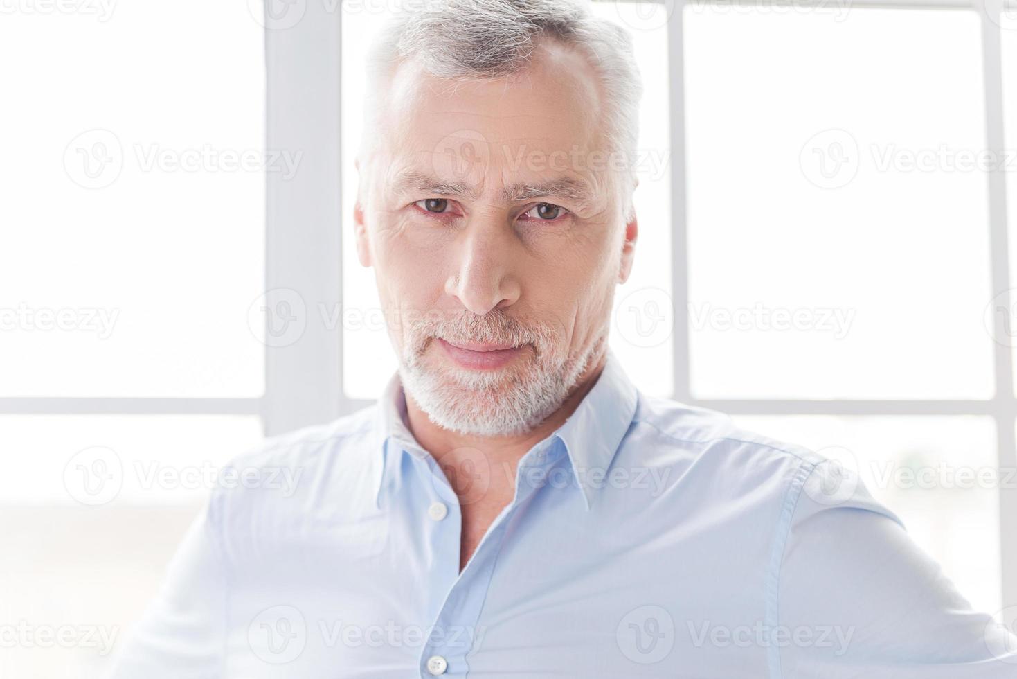 confiado y exitoso. hombre mayor de pelo gris confiado en camisa mirando a la cámara mientras está de pie frente a la ventana foto