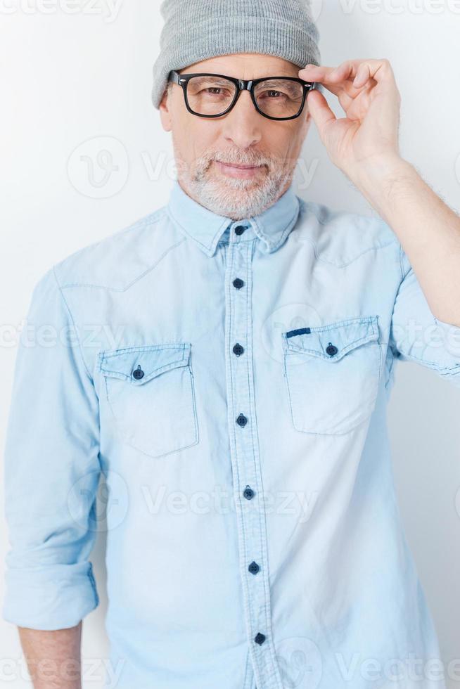 siempre con estilo. hombre mayor de pelo gris confiado en un sombrero funky ajustando sus anteojos y sonriendo mientras está de pie contra el fondo blanco foto