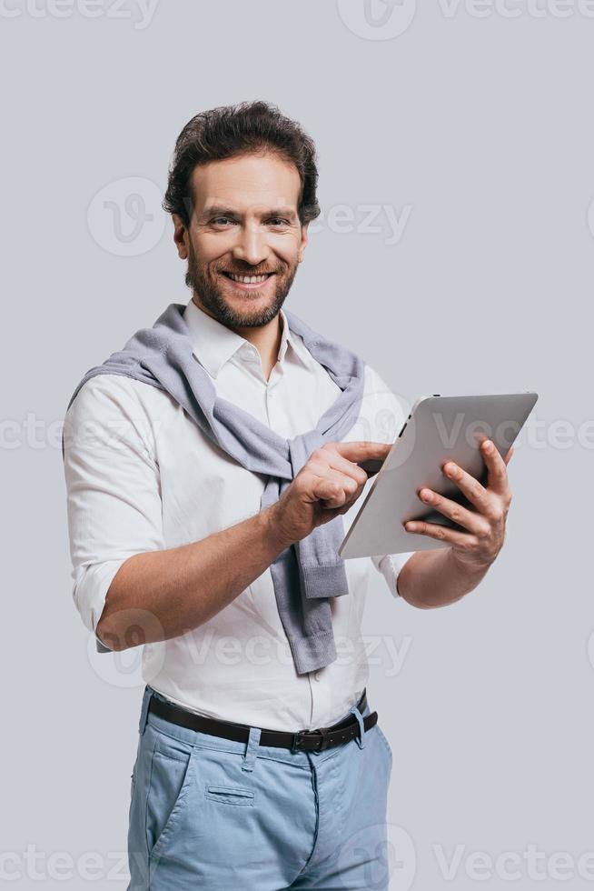 Using modern technologies. Beautiful young man in smart casual clothes working on digital tablet and looking at camera while standing against grey background photo