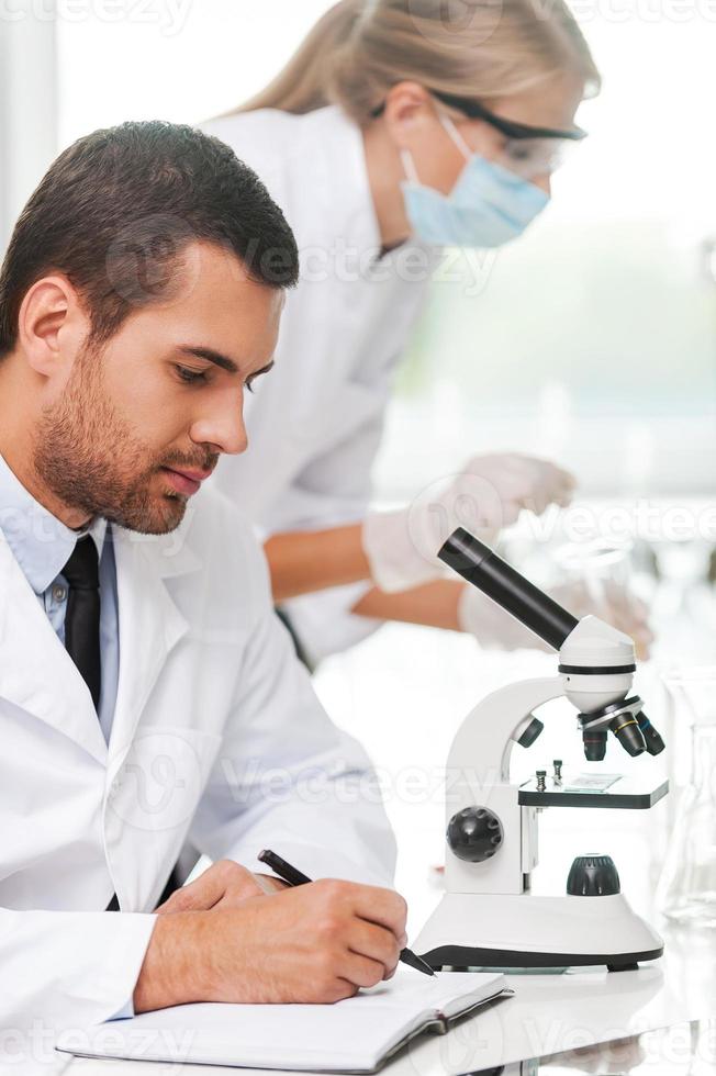 Our goal is to find the cure. Side view of male scientist writing in note pad while his female colleague making experiment in the background photo