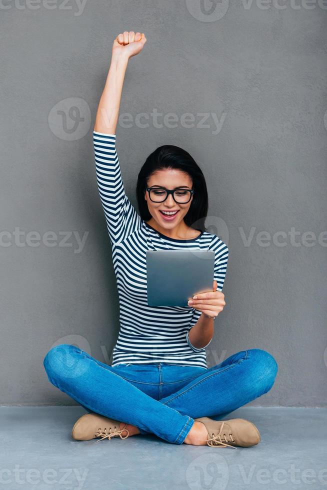 I did it Happy young woman holding digital tablet and keeping arm raised while sitting on the floor and against grey background photo