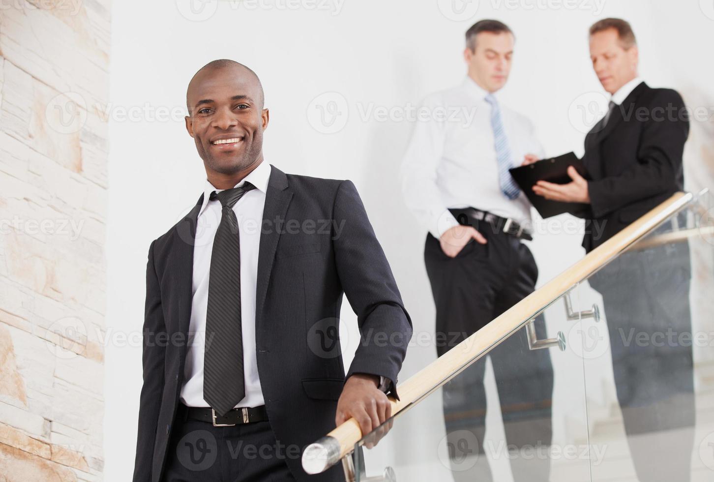 hombre de negocios confiado. alegre joven negro con ropa formal bajando por las escaleras y sonriendo mientras dos personas hablan de fondo foto