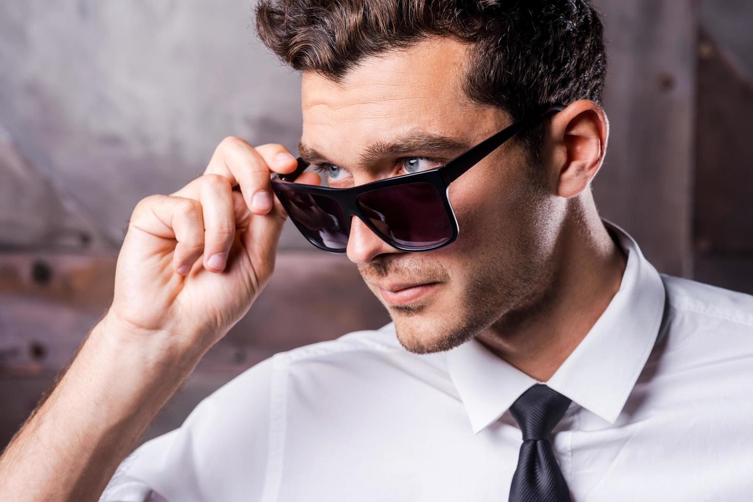 Cool and handsome. Confident young man in shirt and tie adjusting his sunglasses and looking away while standing against metal background photo