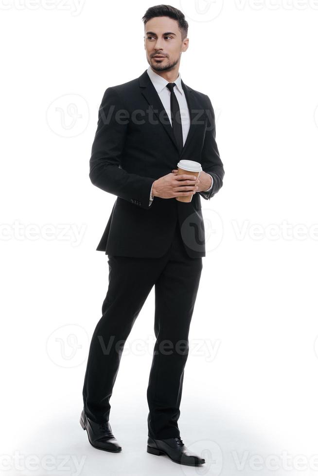 Confidence in every move. Full length of handsome young man in full suit holding disposable cup and looking away while standing against white background photo