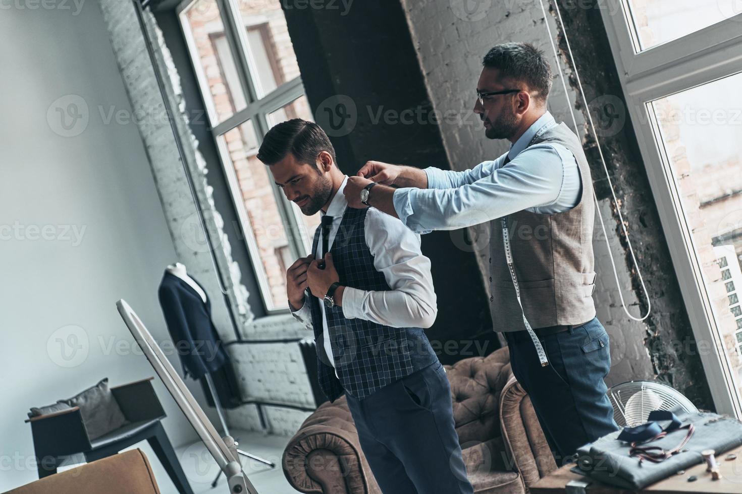 Thinking about every detail. Young fashionable designer helping his client to get dressed while standing in his workshop photo