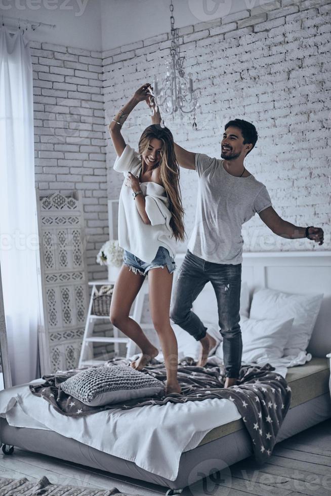 Enjoying every minute with each other. Full length of playful young people holding hands and smiling while dancing on the bed at home photo