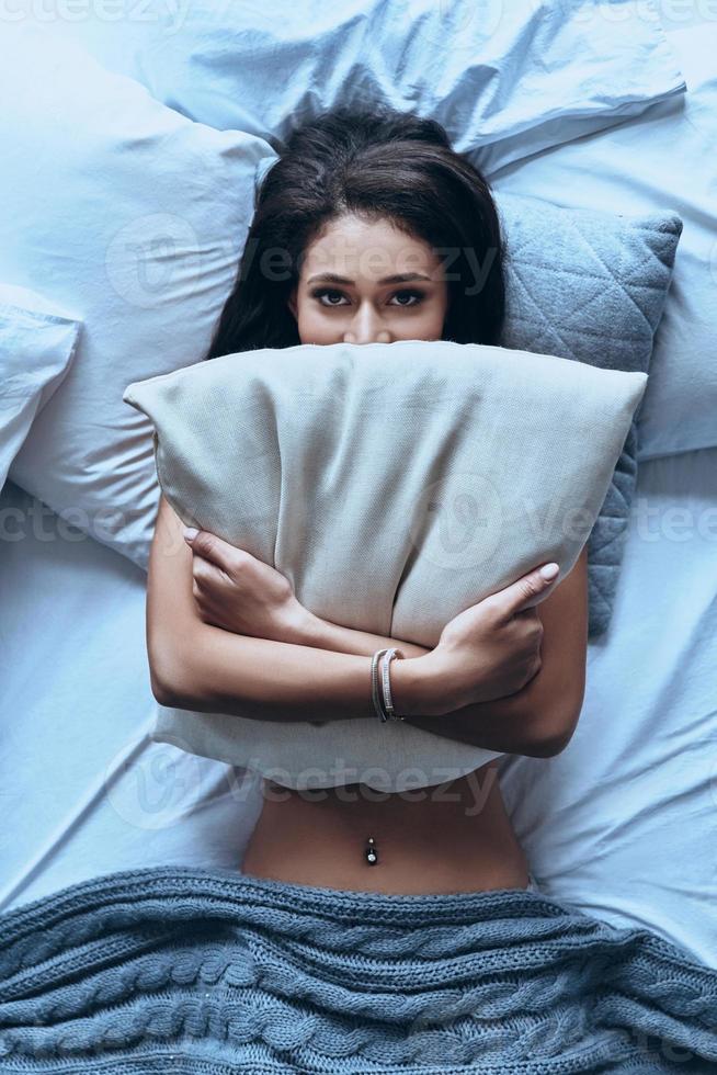 Just one more minute in bed Top view of attractive young woman covering half of her face with pillow and looking at camera while lying on the bed at home photo