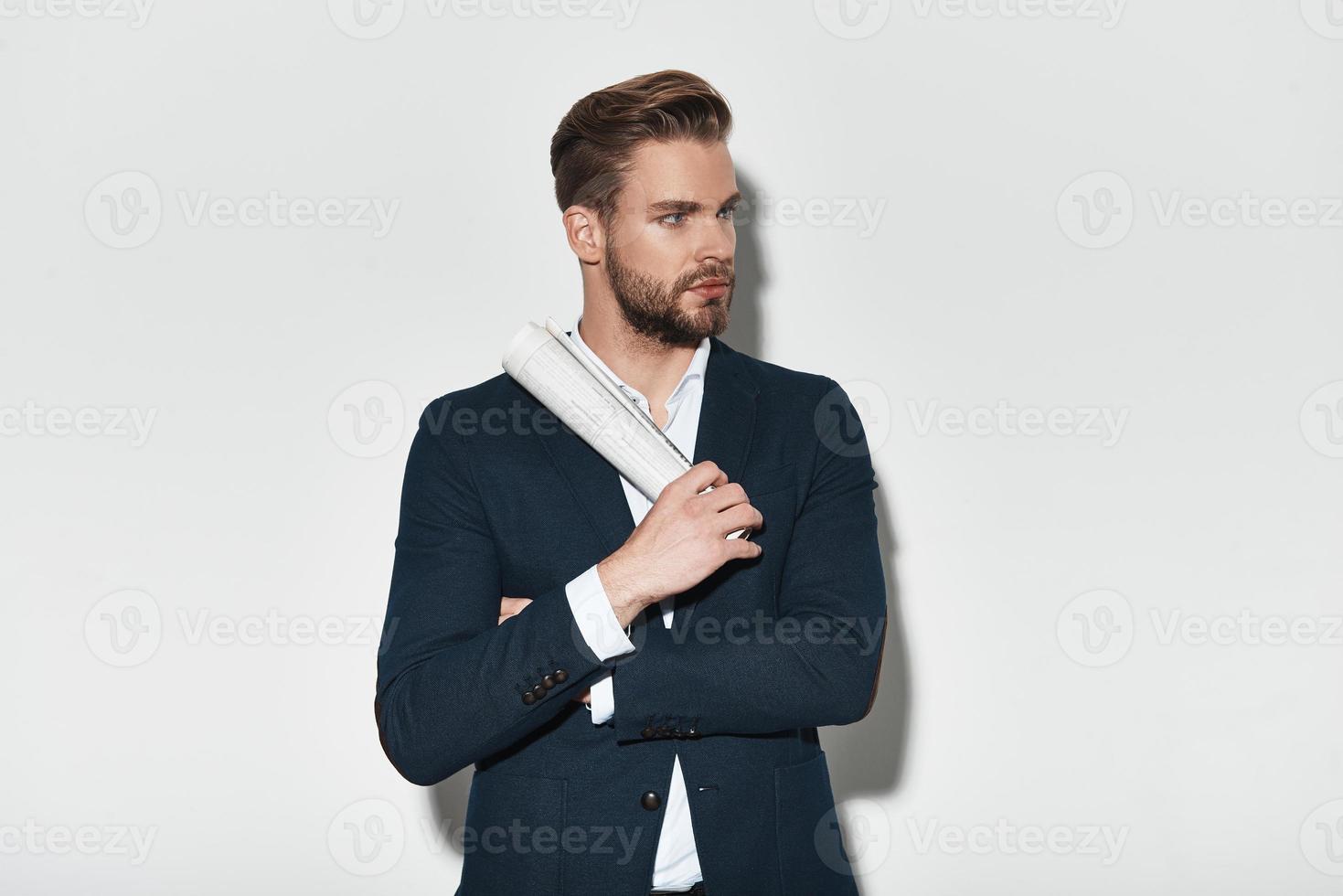 Confidence and charisma. Handsome young man in full suit keeping arms crossed and looking away while standing against grey background photo