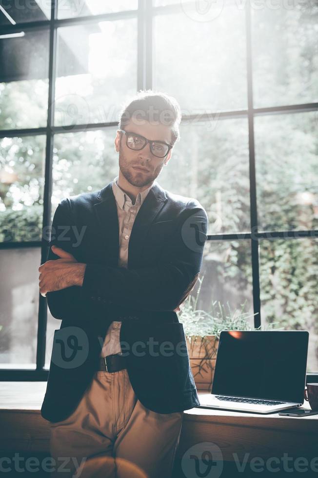 Young and successful. Confident young man in smart casual wear keeping arms crossed and looking at camera while standing in front of the big window in office or cafe photo