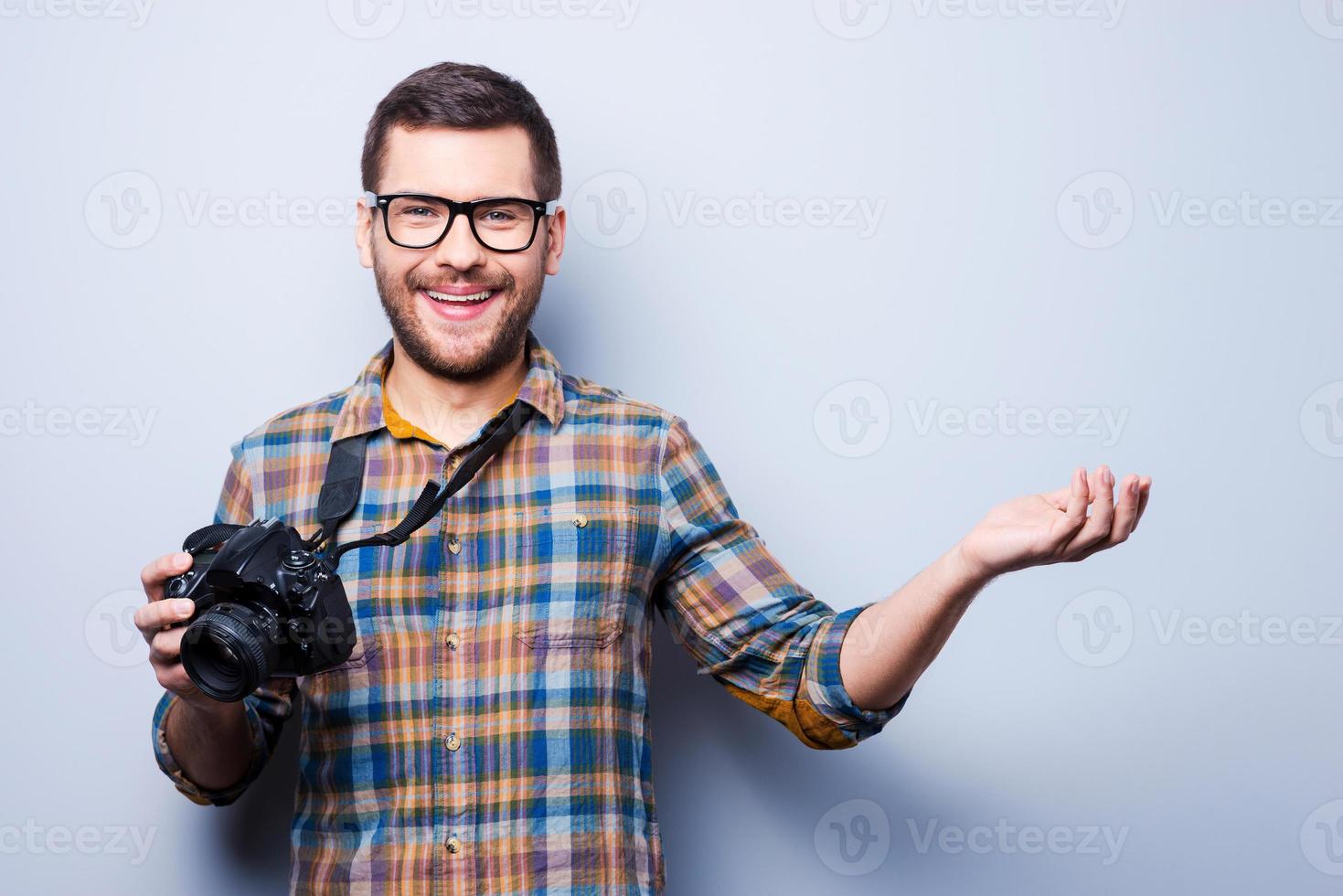 ven a mí para obtener una buena foto. retrato de un joven confiado en camisa sosteniendo la cámara y señalando mientras está de pie contra un fondo gris foto