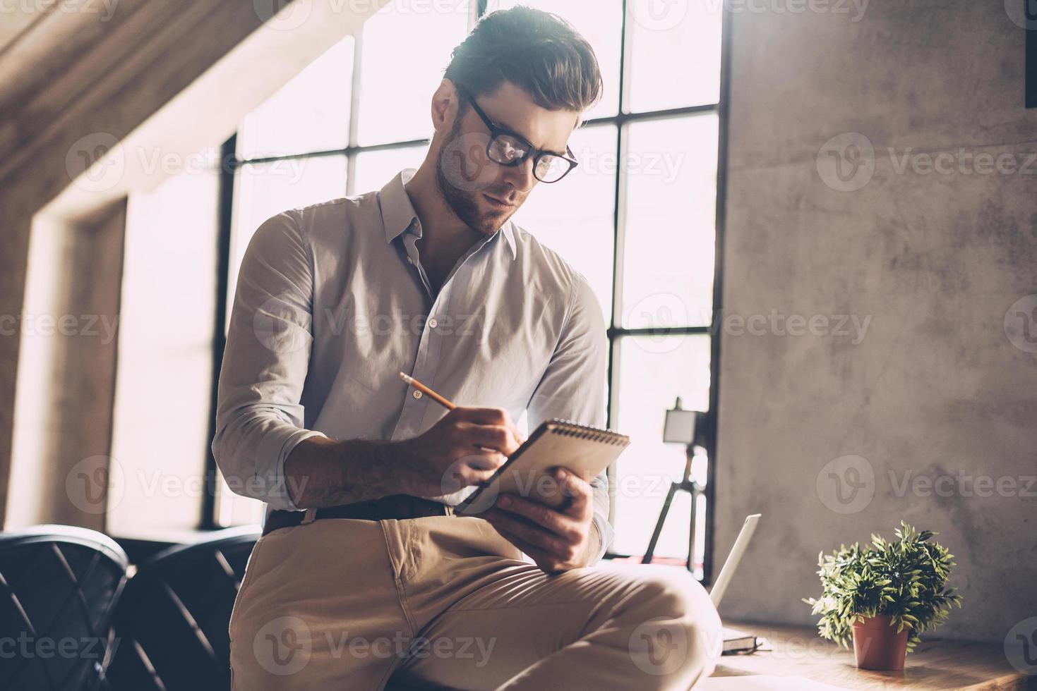 Just inspired. Confident young man in smart casual wear writing something in his notebook while leaning at the desk in office photo