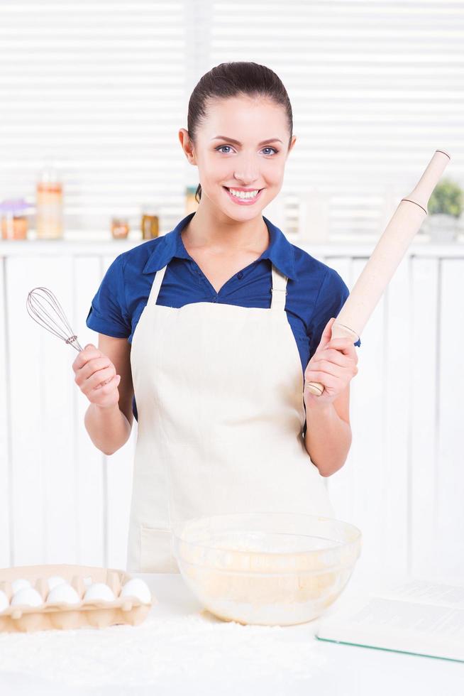 Ready for cooking. Cheerful young woman in apron holding rolling pin and wire whisk while standing in a kitchen photo