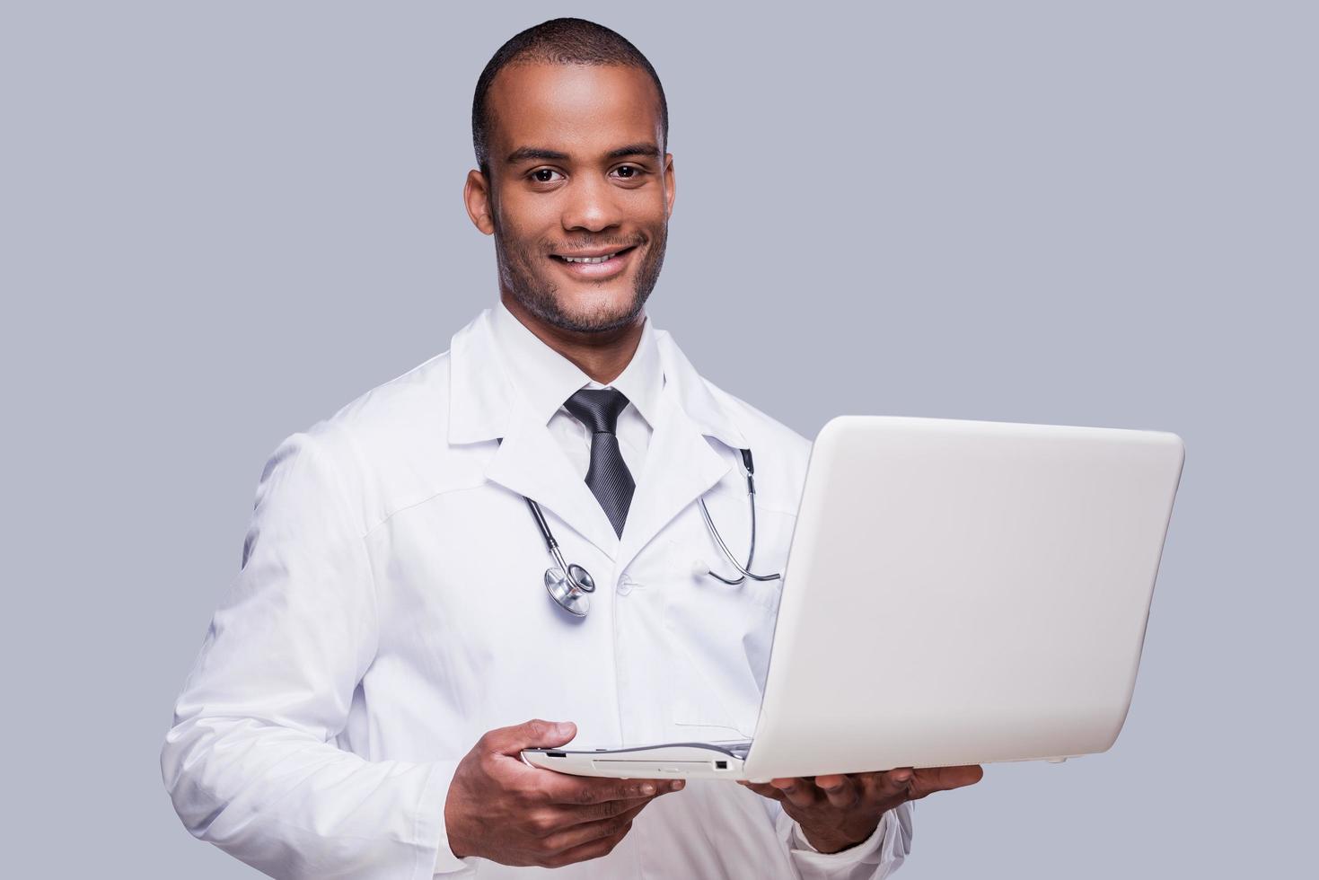 Confident medical doctor. Confident African doctor holding laptop and smiling while standing against grey background photo