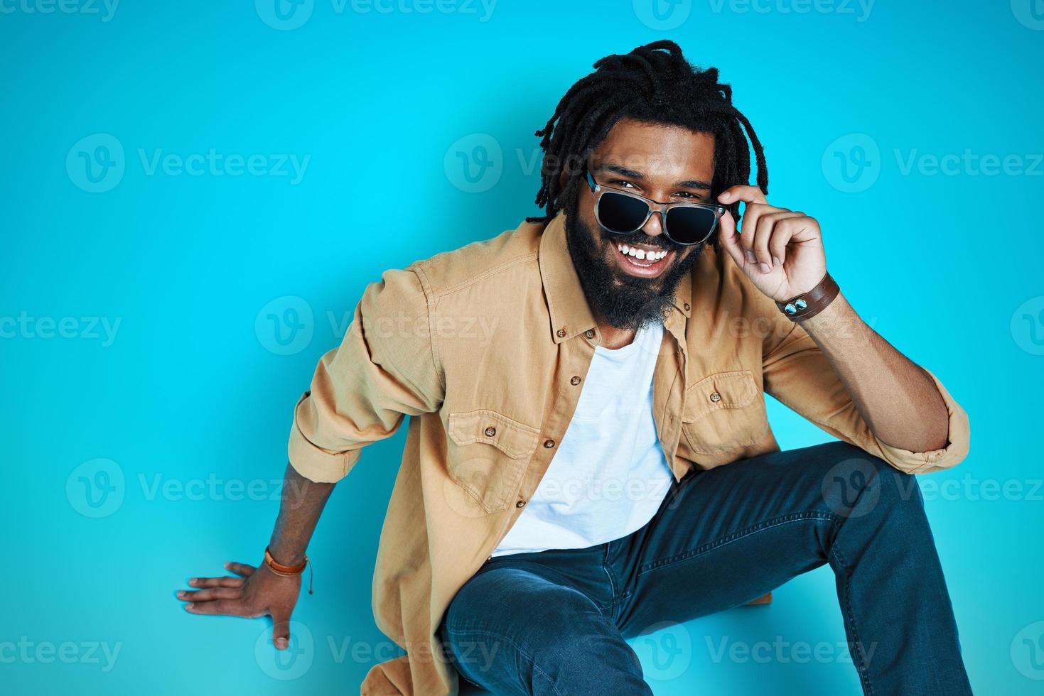 Top view of smiling young African man in casual wear adjusting eyewear and looking at camera while sitting against blue background photo