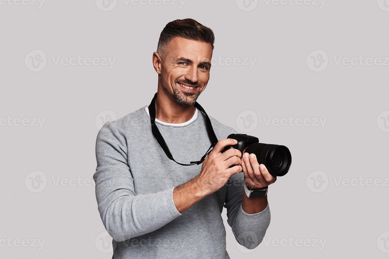 His best hobby. Good looking young man holding digital camera and smiling while standing against grey background photo